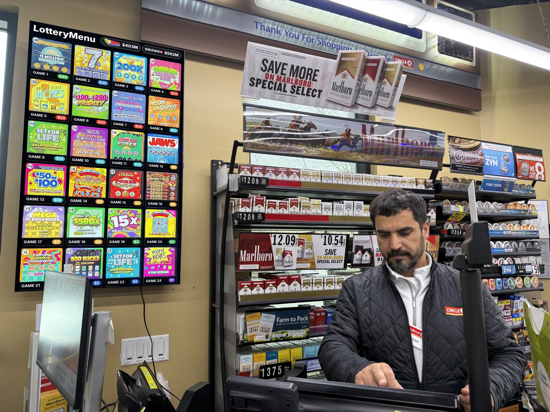 Cashier at a convenience store checkout counter beneath a wall-mounted LotteryMenu board showing 24 scratch-off games and jackpot amounts